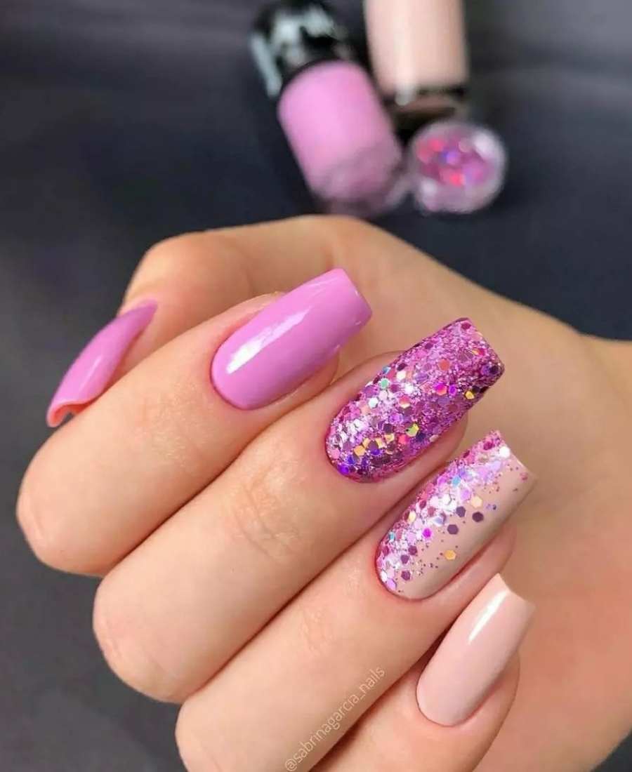 Uma mão com unhas pintadas, polegas e indicador de rosa, mínimo e anelar de nude, o dedo médio está com glitter pink e o dedo anelar com um pouco também