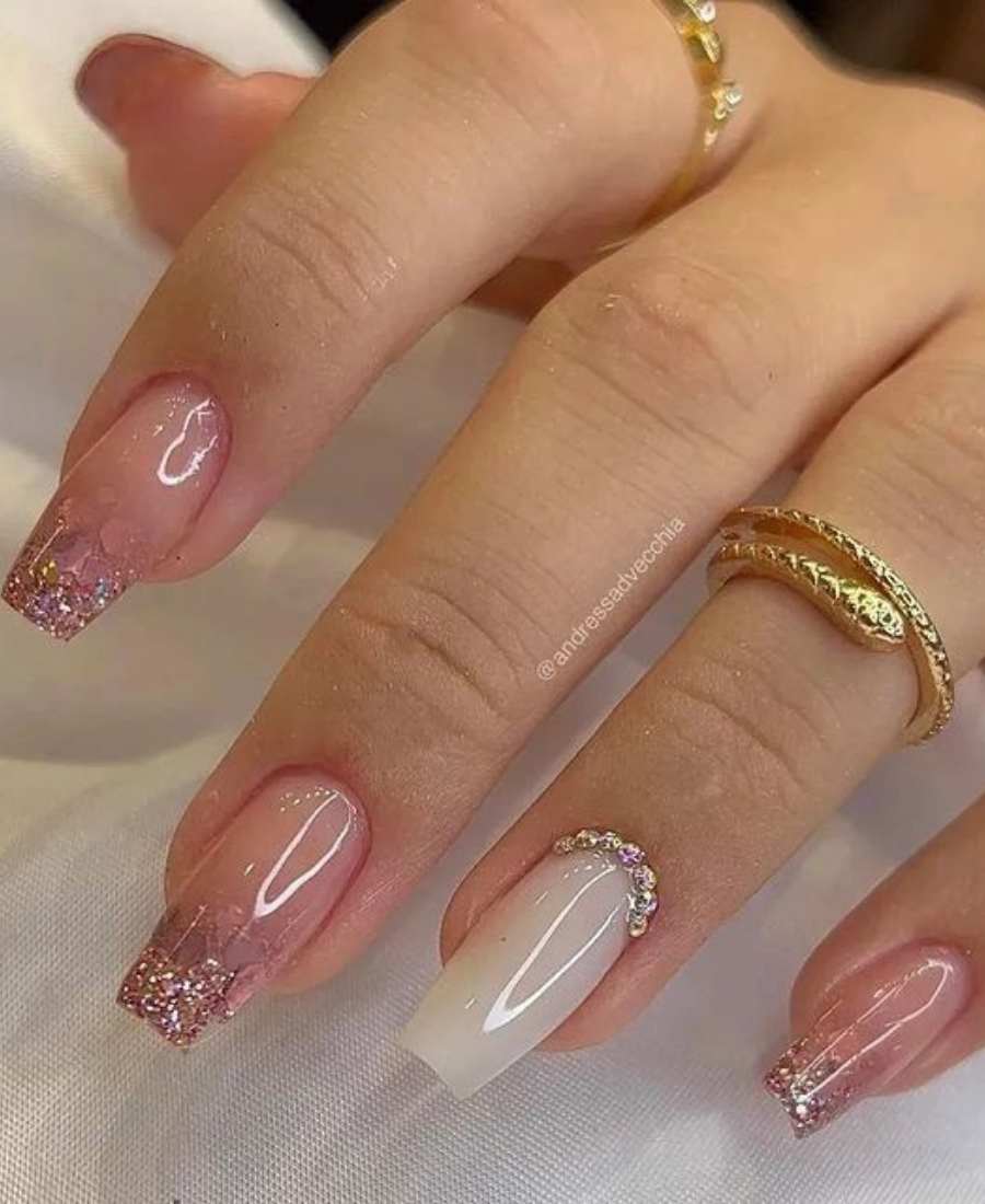 Mão decorada. Dedos mínimo, médio e indicador estão pintados de rosa-claro com glitter na ponta, enquanto o dedo anelar está branco com pedrarias na base.