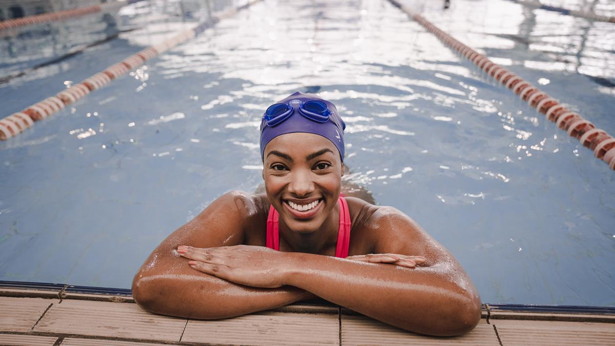 Mulher apoiada na borda da piscina usando maiô vermelho e touca de natação azul da Oxer