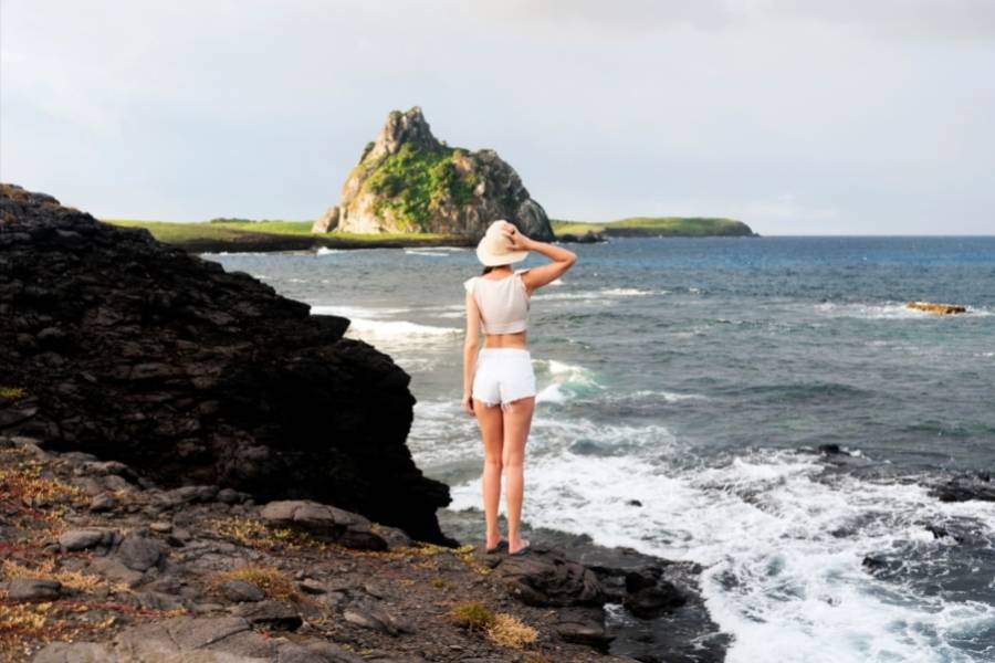 mulher de costas na praia usadndo chapéu, cropped e shorts branco