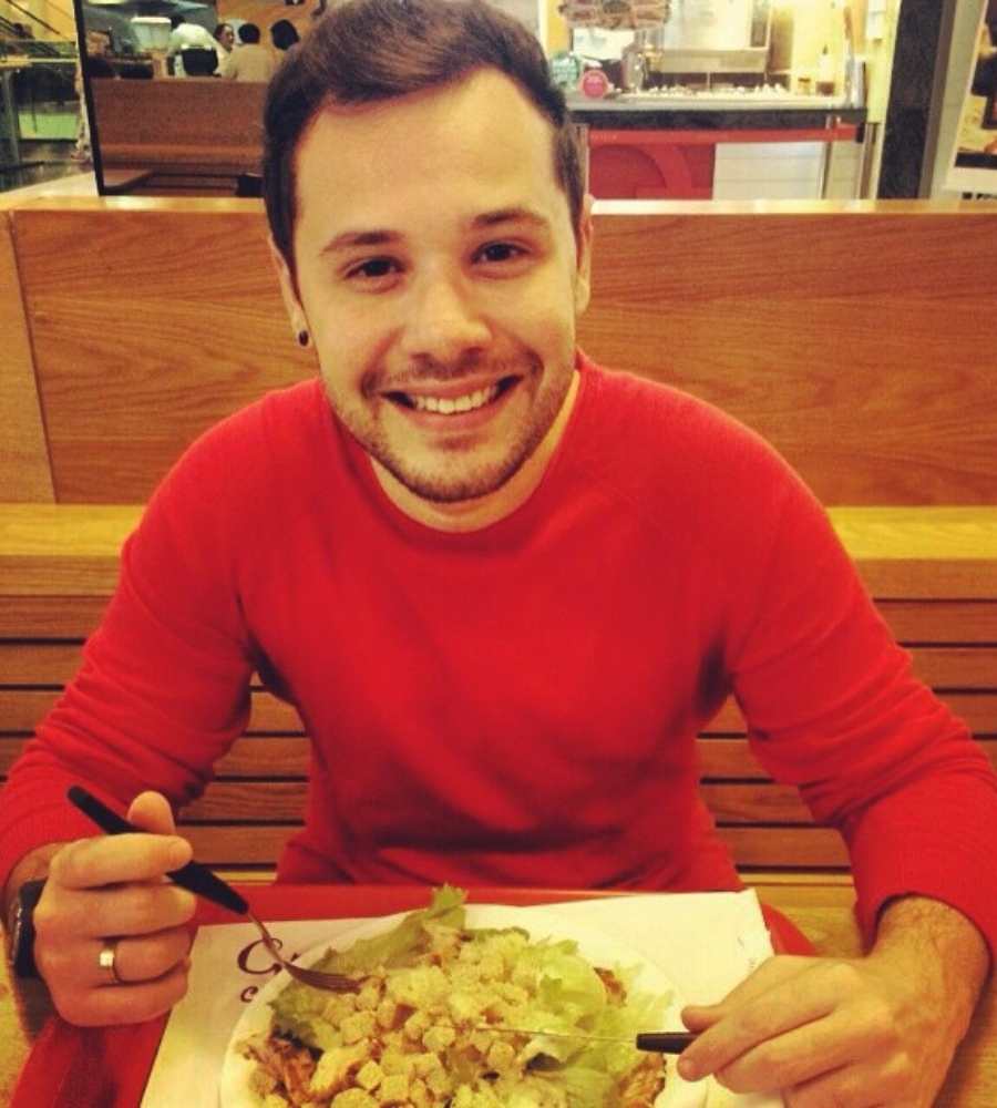 Imagem em fundo de áre de refeição de restaurant. No centro, sentado em banco, ex-A Casa segurando garfo com prato de salada na mesa. Ele sorri e veste camisa de frio vermelha. 