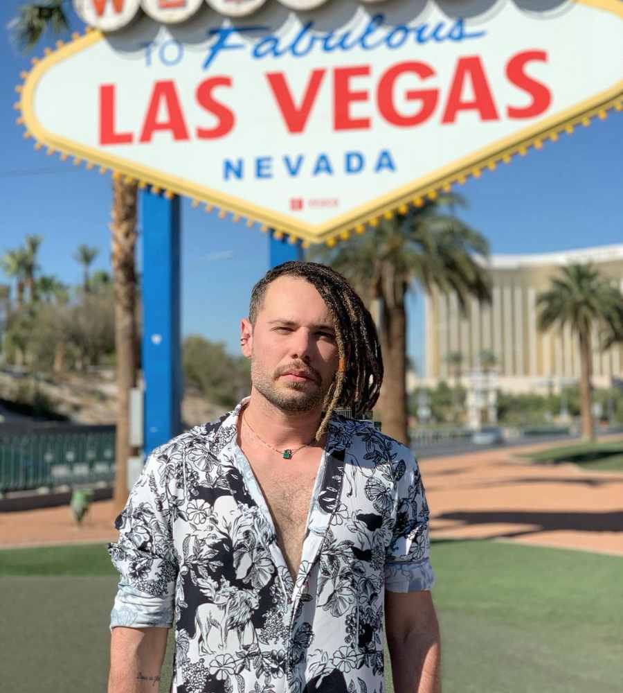 Imagem em fundo da placa de Las Vegas, com gramado, construções e palmeiras no fundo. No centro, Alex Gallete de dreads usando camiseta estampa preta e branca.
