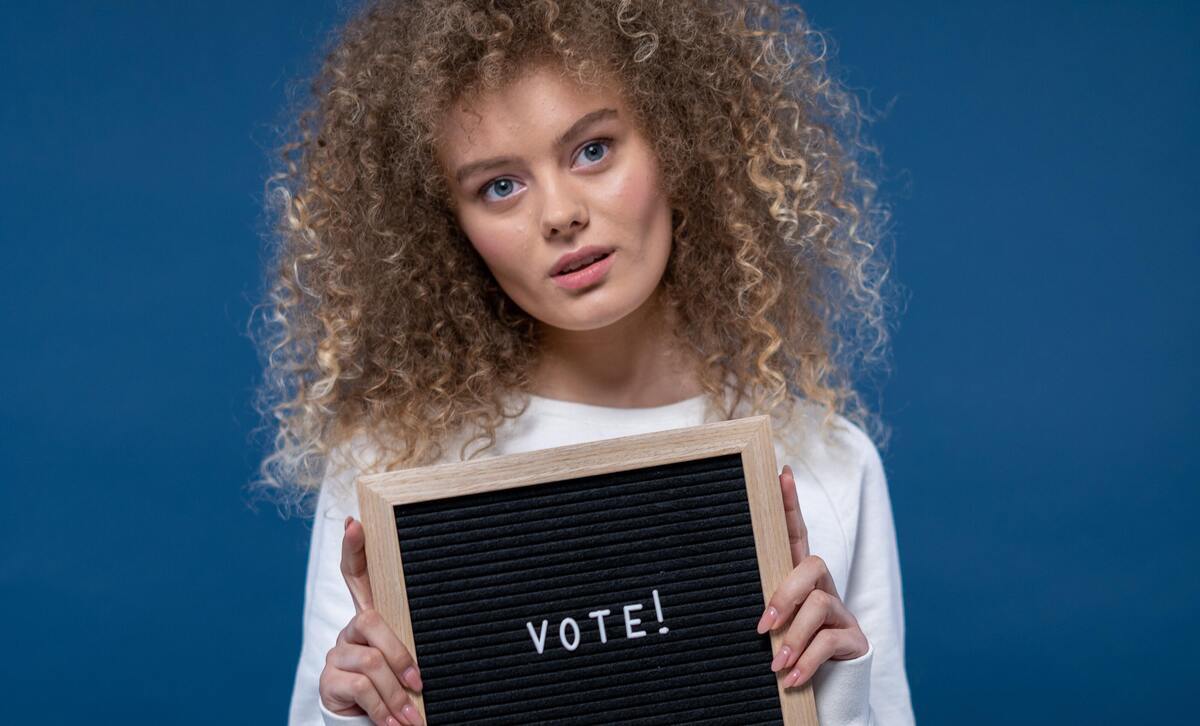mulher loira de olhos claros com cabelos cacheados segurando uma placa escrito "vote" para representar o eleitor de cada signo