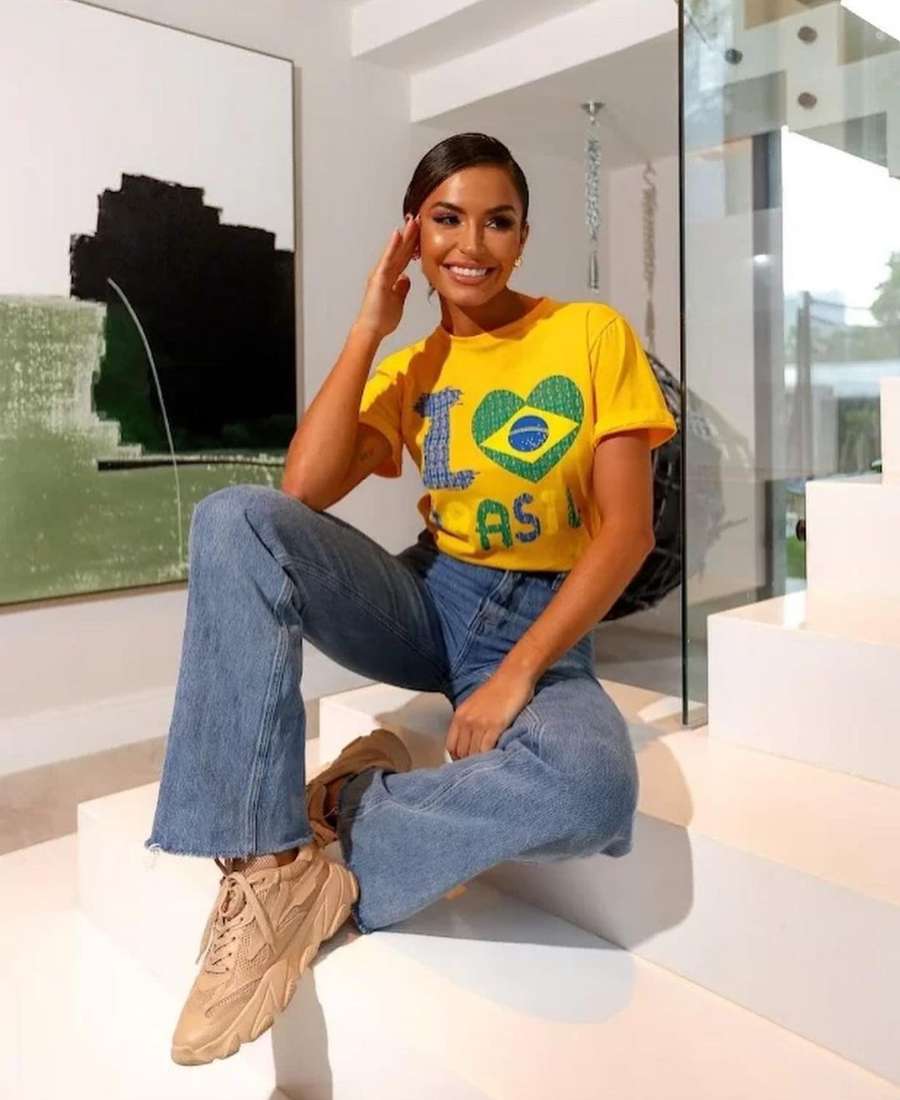 Imagem com fundo de cômodo de casa, com parede e escada. No centro, mulher usando roupas para a copa do mundo: camisa amarela escrito "I love Brasil", calça jeans e tênis bege.