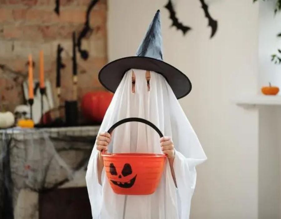 Criança fantasiada de fantasma com chapéu de bruxa segurando balde de abóbora