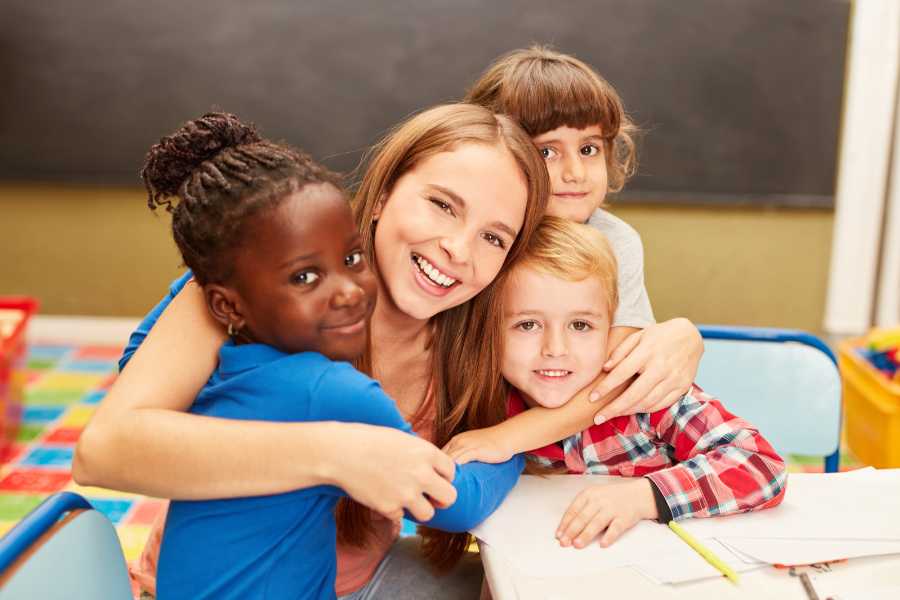 Foto de professora abraçada com alunos. Todos estão sentados em mesa na sala de aula.