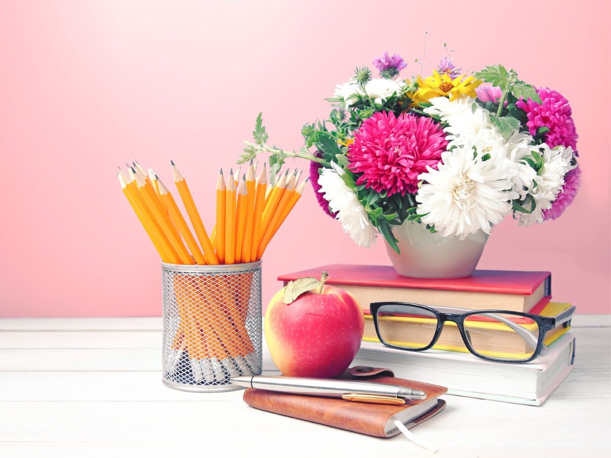 Foto de livro, óculos, maça, agenda, porta lápis com lápis e pote com flores, tudo sobre superfície branca.