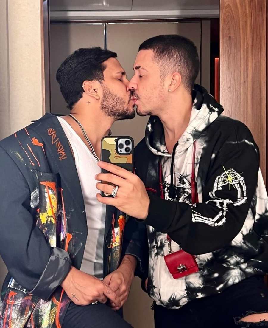 Foto de Carlinhos Maia e Lucas Guimarães se beijando em frente a espelho em selfie. Os dois vestem roupa de frio estilosa: blazer e moletom coloridos.