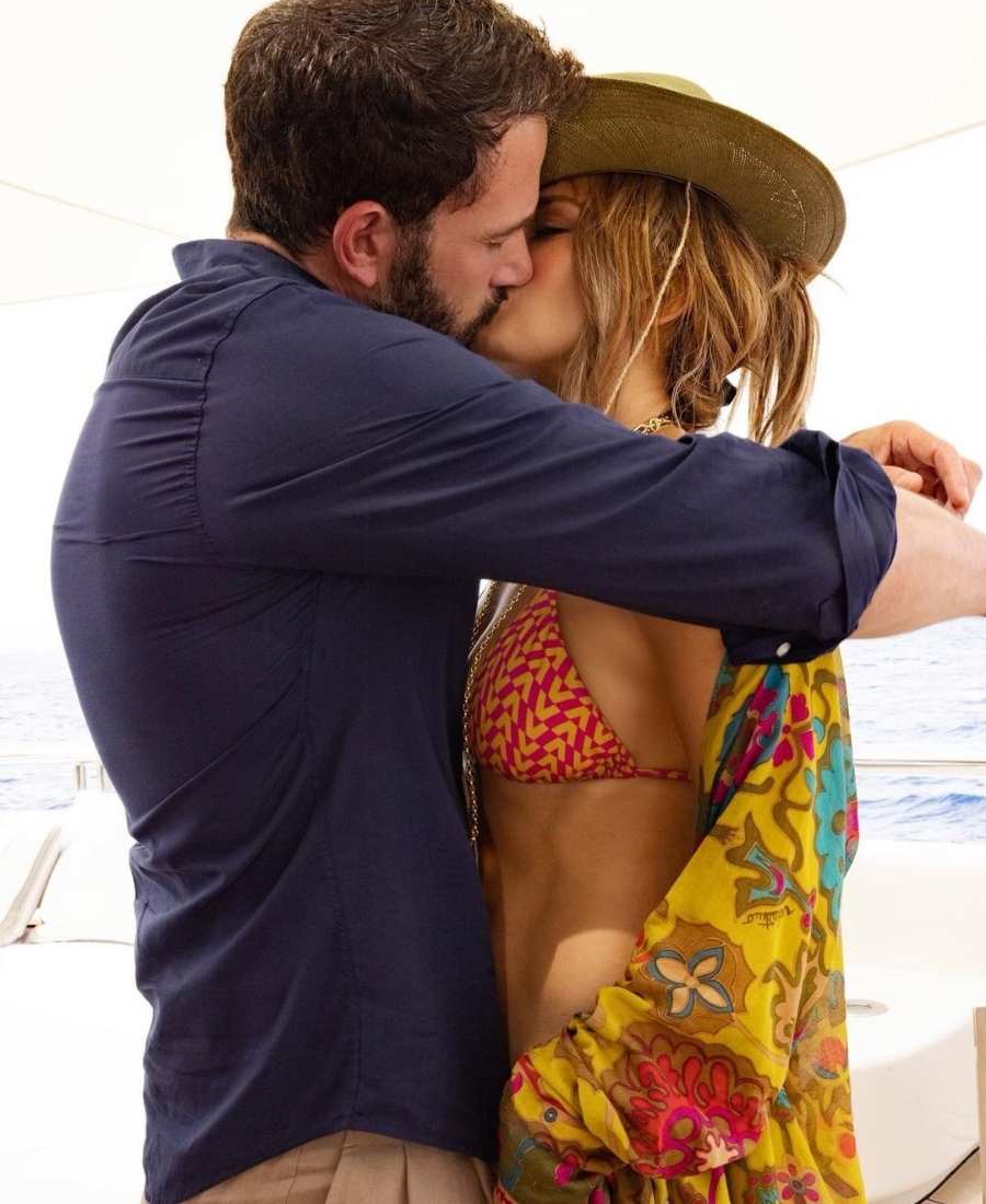 Imagem com fundo de mar, com Jennifer Lopes e Ben Affleck se beijando em embarcação. Ela veste biquíni vermelho e amarelo minúsculo. Ele camisa azul. 