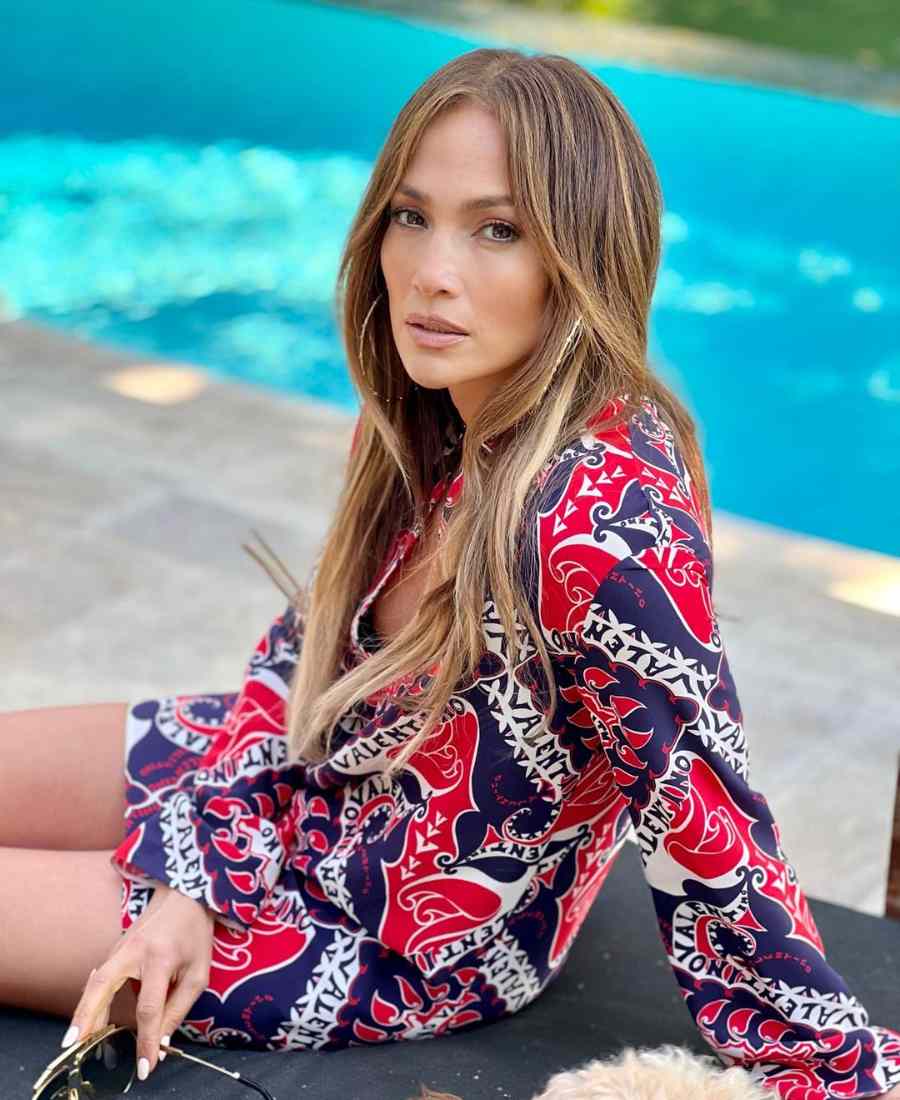 Imagem com fundo de piscina. No centro, sentada no chão, Jennifer Lopez usando vestido da Valentino estampado de cor azul, vermelho e branco. Ela usa brincos de argola e segura óculos de sol.