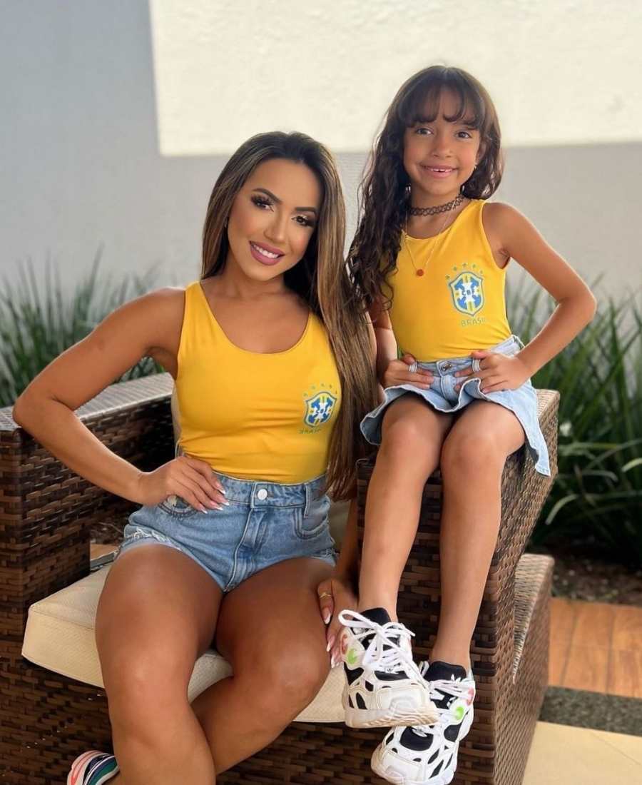 Imagem com fundo de área esterna de casa. No centro, mulher e menina criança usando roupa para Copa do Mundo: ambas usam body amarelo, a adulta de short jeans e a pequena de saia jeans. 