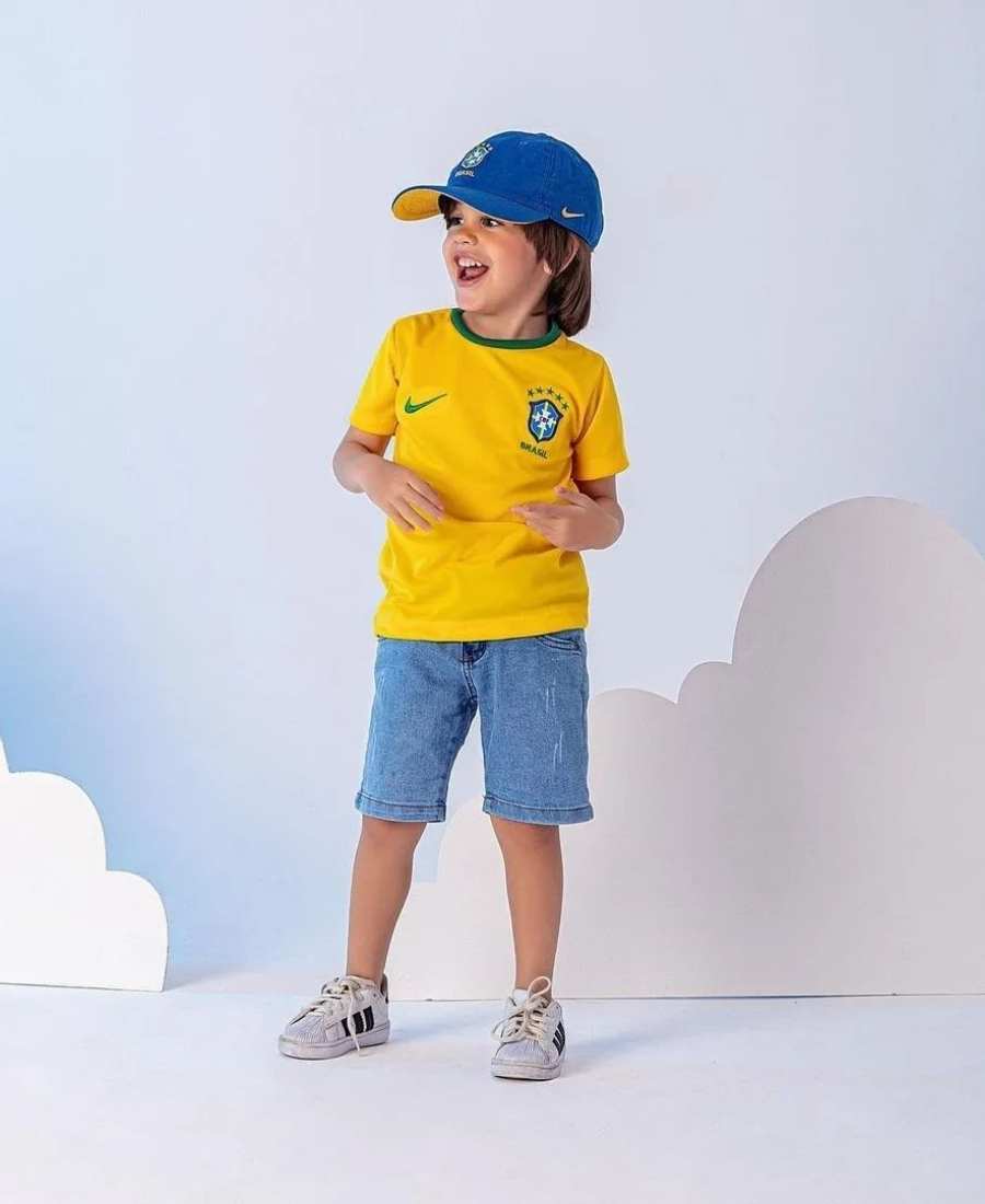 Imagem com fundo branco com nuvens. No centro, menino criança usando roupa Brazilcore: camiseta amarela do Brasil + bermuda jeans + boné azul do Brasil + tênis branco.
