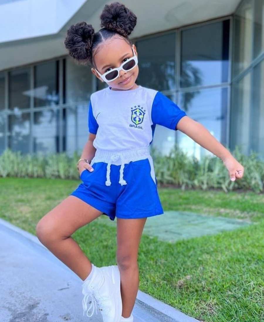 Imagem com fundo de área esterna de casa. No centro, menina criança usando roupa para Copa do Mundo: camiseta branca e azul do Brasil + short branco e azul + tênis branco.