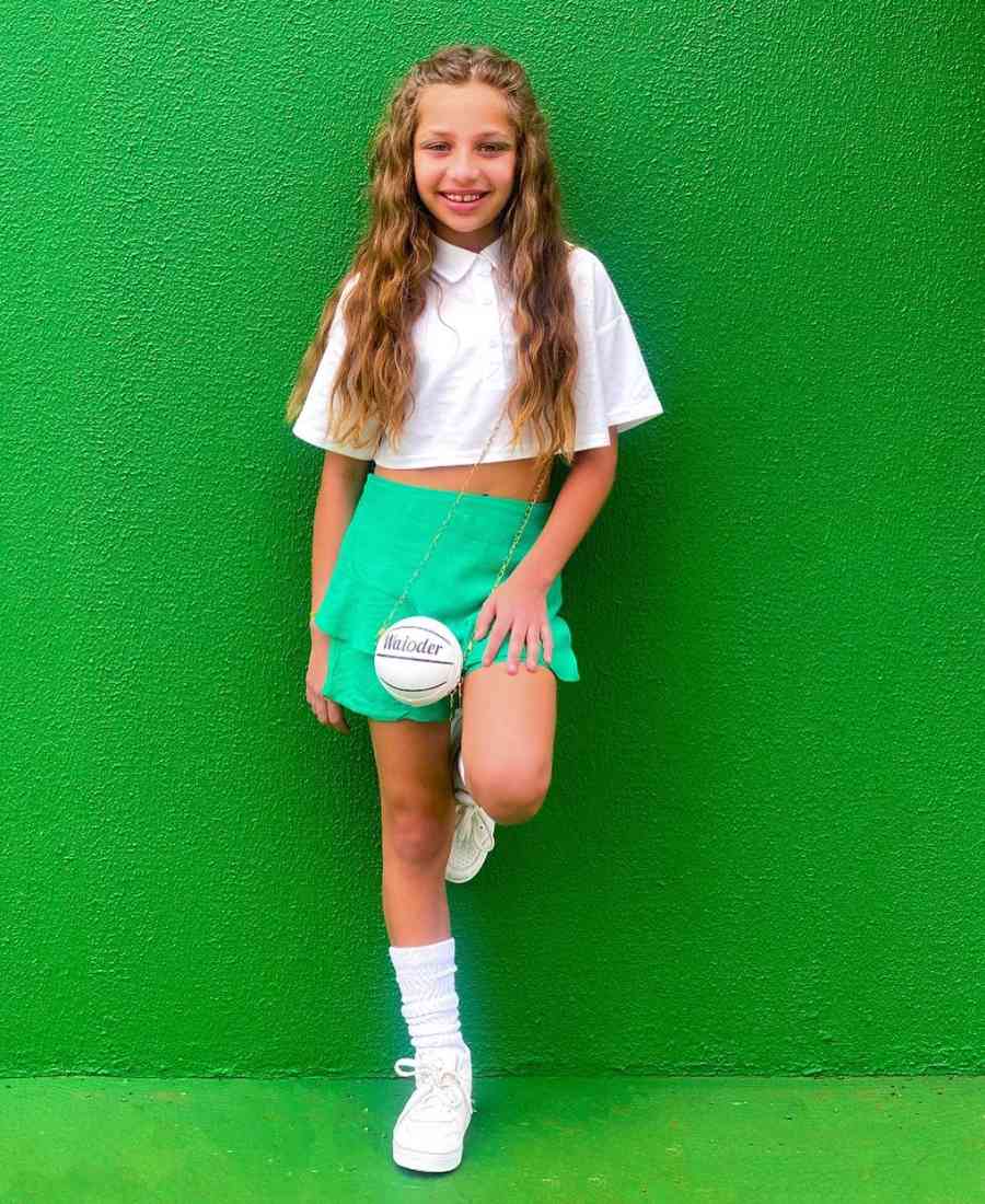 Imagem com fundo de parede verde. No centro, menina criança usando roupa para Copa do Mundo: cropped branco + saia verde + tênis branco + bolsa de bola. 