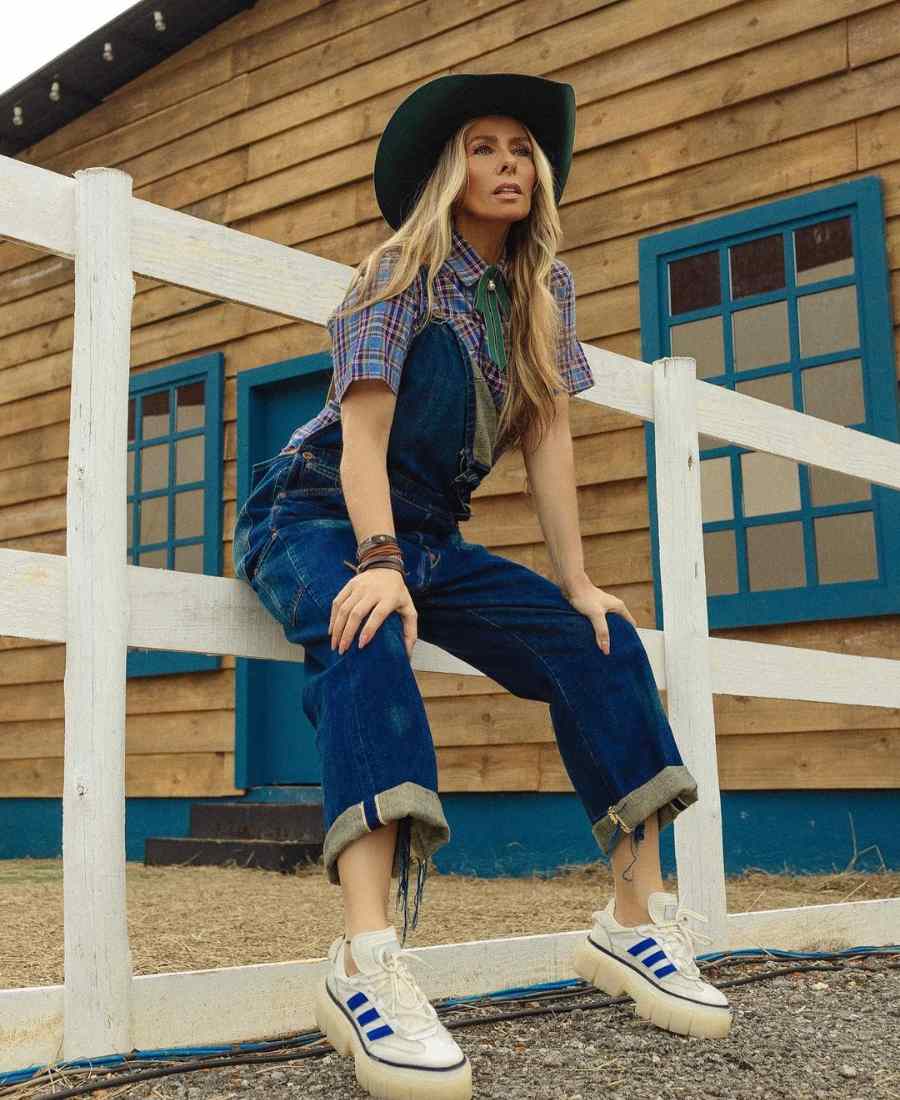 Imagem de Adriane Galisteu com look para apresentar A Fazenda. Ela está sentada em cerca branca de casa de madeira com janelas azuis. 