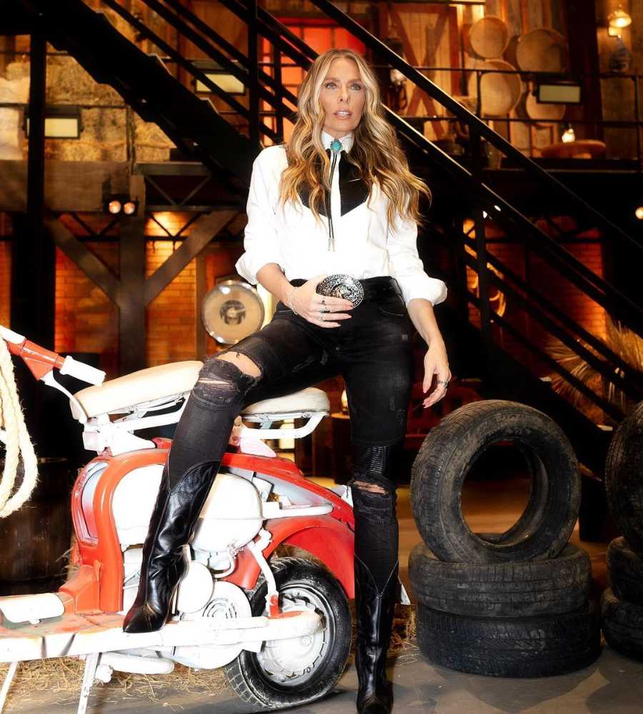 Imagem em fundo do estúdio do A Fazenda, com pneus velho, motocicleta, escada, feno, etc. No centro, Adriane Galisteu apoiando pé na moto e segurando fivela do cinto com uma das mãos.