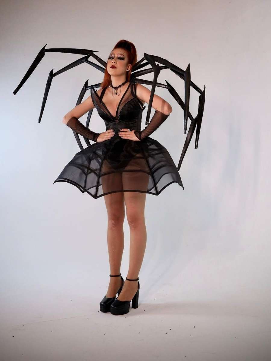 Imagem em fundo branco com foto de Camila Pudim no centro. Ela está usando fantasia de aranha. A peça é um vestido preto e na parte de traz do pescoço traz as pernas grandes da aranha.
