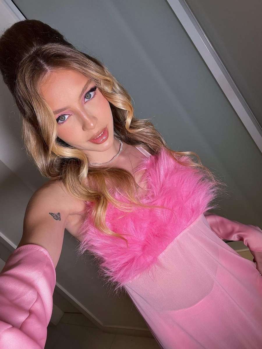 Imagem em fundo claro com parede. Na frente, em uma selfie, Fefe fantasiada para o Halloween Sephora de personagem do filme "Austin Powers" (1997). Ela veste vestido transparente pink com plumas no decote e luvas.