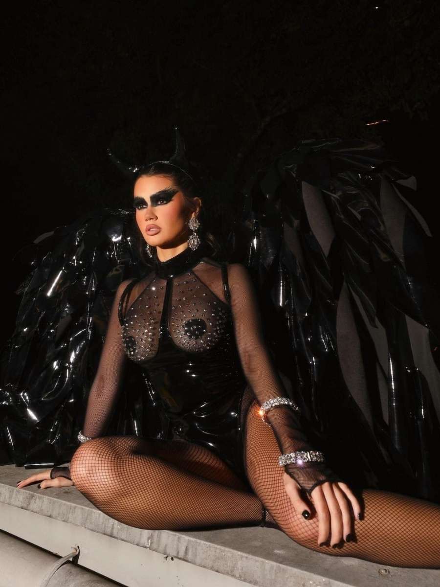 Imagem em fundo preto com Franciny sentada em superfície de cimento. Ela está fantasiada para o Halloween Sephora de anjo negro. Usa body preto de vinil com meia calça e segunda pele arrastão, tiara de chifres e asas gigantes pretas.
