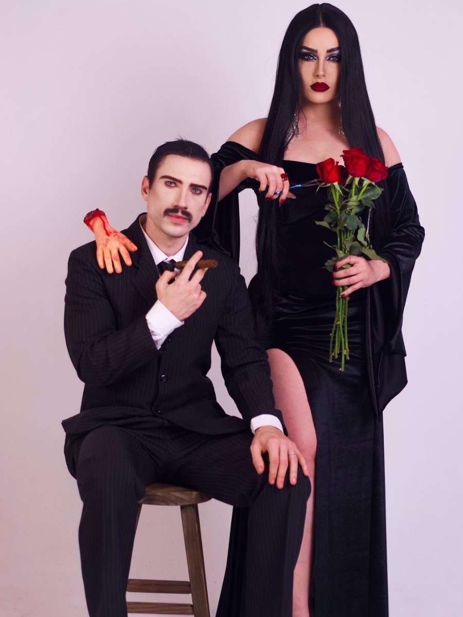 Imagem em fundo rosa-claro com Jessica Cardoso e Dreicon usando fantasia inspirada no filme "A Família Addams". Ele está usando um terno preto, segurando charuto e sentado em banco de madeira. Ela, usa vestido longo de fenda preto, segura rosas-vermelhas e tesoura nas mãos.