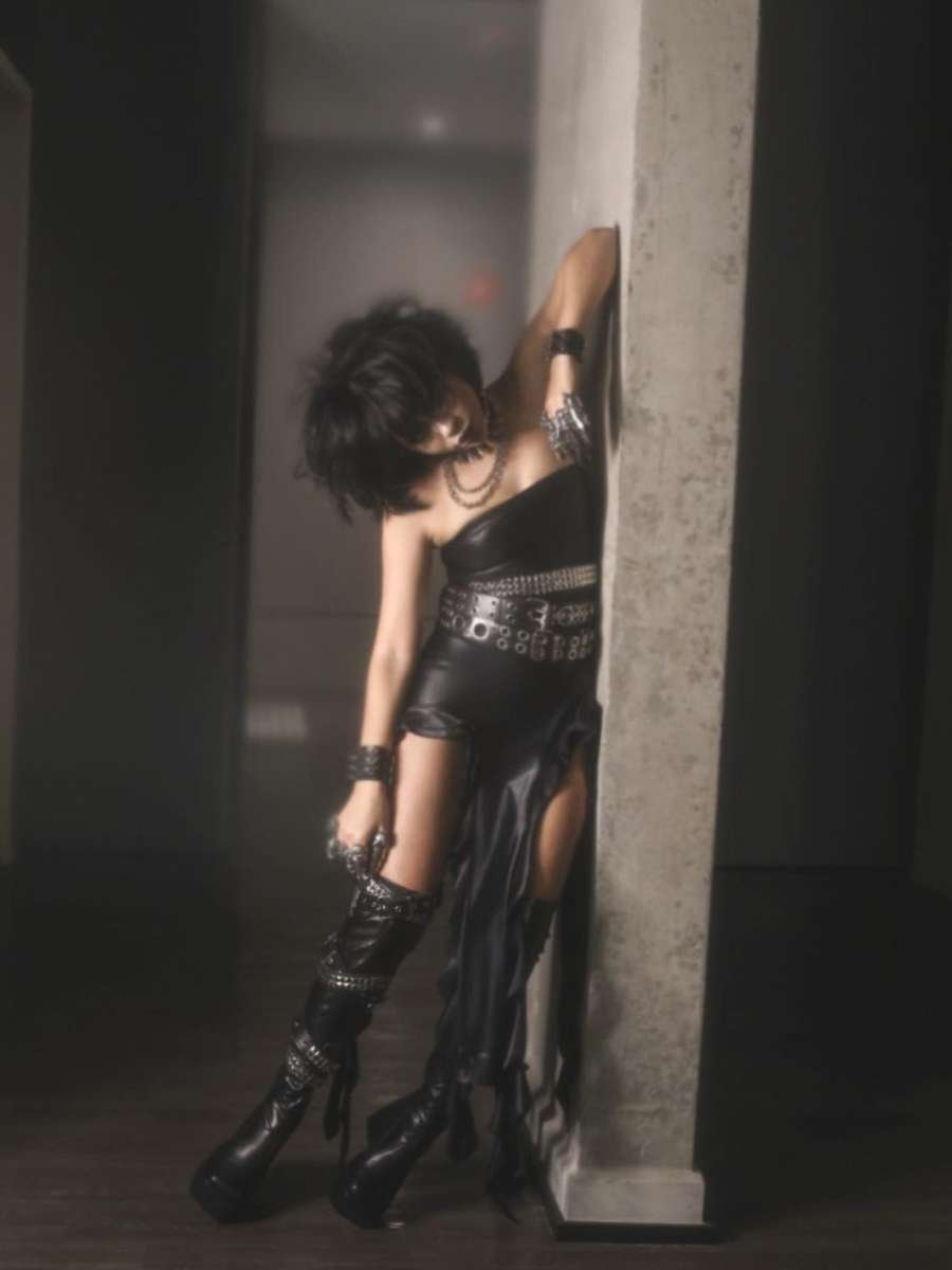Imagem em fundo cinza com parede e Laura Brito encostada nela. Ela está usando fantasia do Edward mãos de tesoura. É um vestido de couro preto com cintos na cintura.