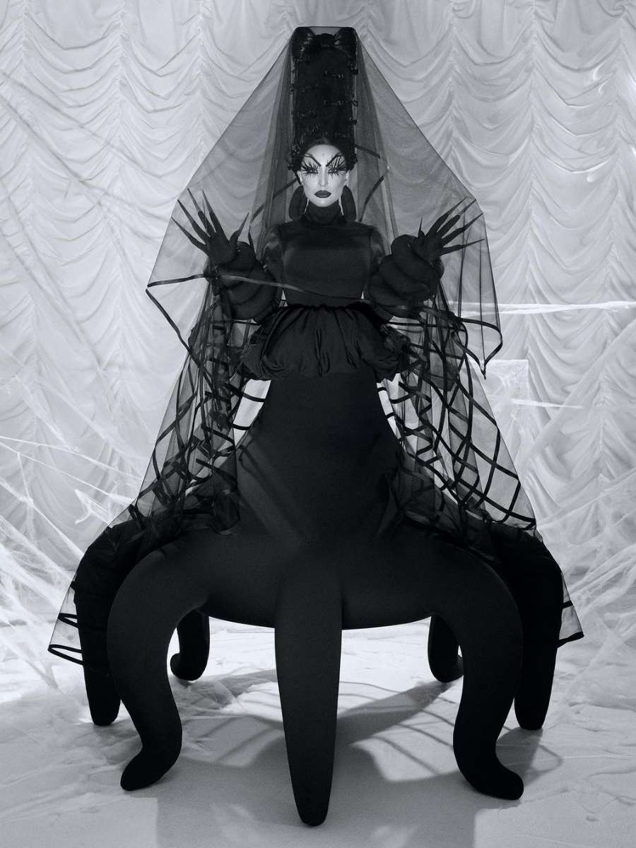 Imagem em preto e branco No centro, Sabrina Sato usando fantasia bizarra de aranha. A peça é um vestido preto com "pernas". Além disso, tem um véu preto ao seu redor. 