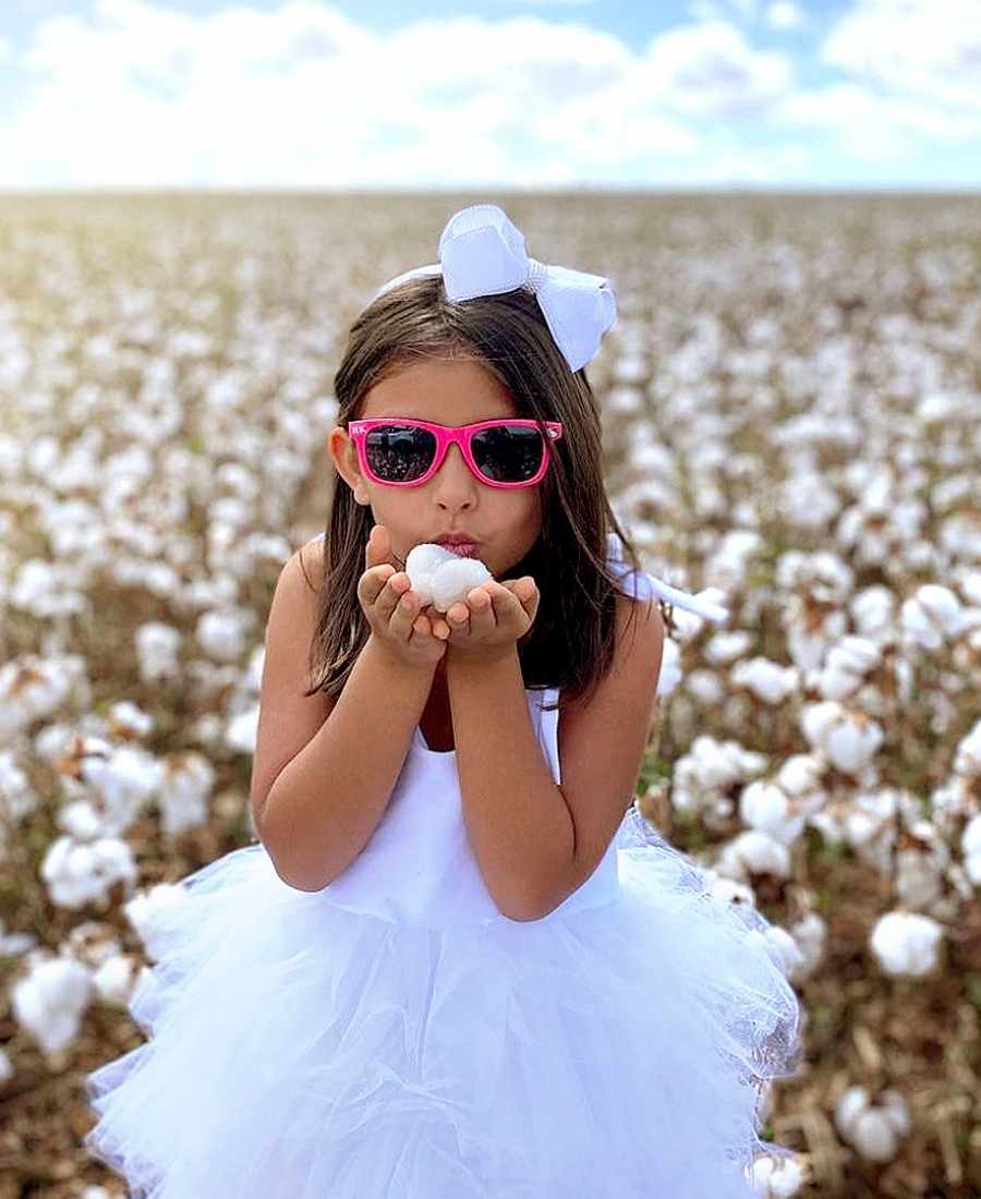 Imagem com fundo de campo de algodão. No centro, menina criança usando vestido branco e óculos rosa, soprando algodão.