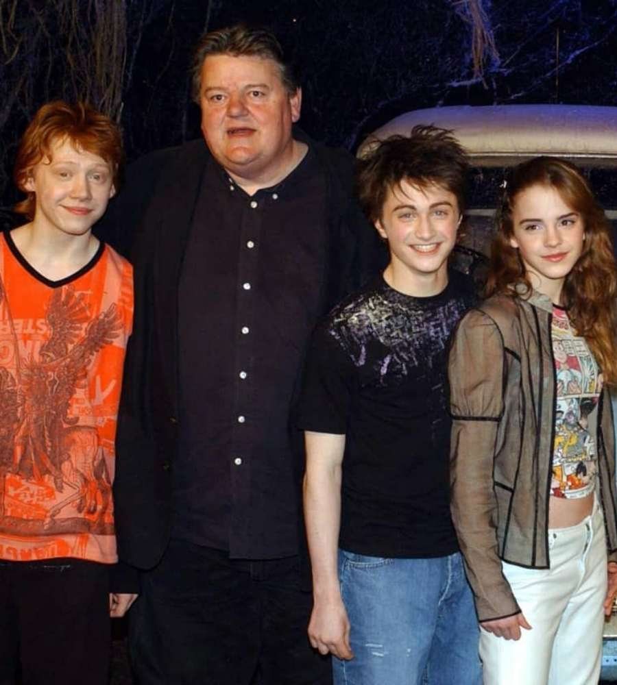 Imagem com fundo preto. Atores Rupert Grint, Robbie Coltrane, Daniel Radcliffe e Emma Watson abraçados.