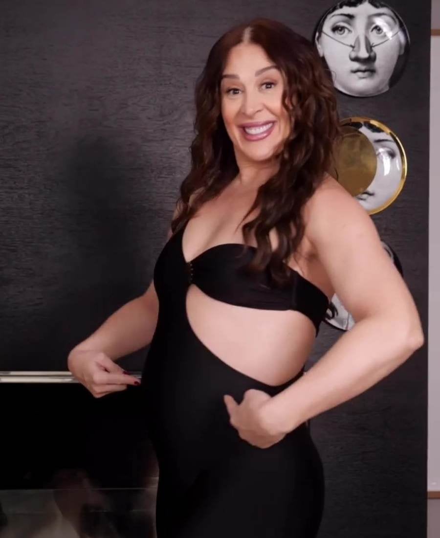 Print de vídeo da atriz anunciando gravidez. Fundo com parede cinza e pratos decorativos. No centro, atriz sorridente apontando os dedos para sua barriga.