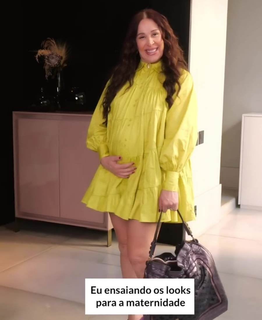 Print de vídeo de Claudia Raia. Imagem com fundo de cômodo de casa, com paredes e móveis. No centro, Claudia Raia sorridente, usando vestido amarelo e segurando malas. Na parte superior da imagem, 