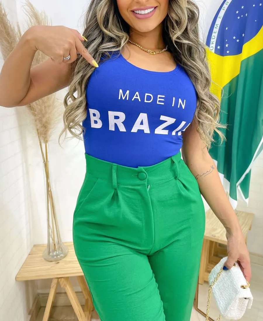 Imagem com fundo de parede, plantas e bandeira do Brasil. No centro, mulher usando roupa copa do mundo: body azul escrito "made in Brazil" + calça verde.