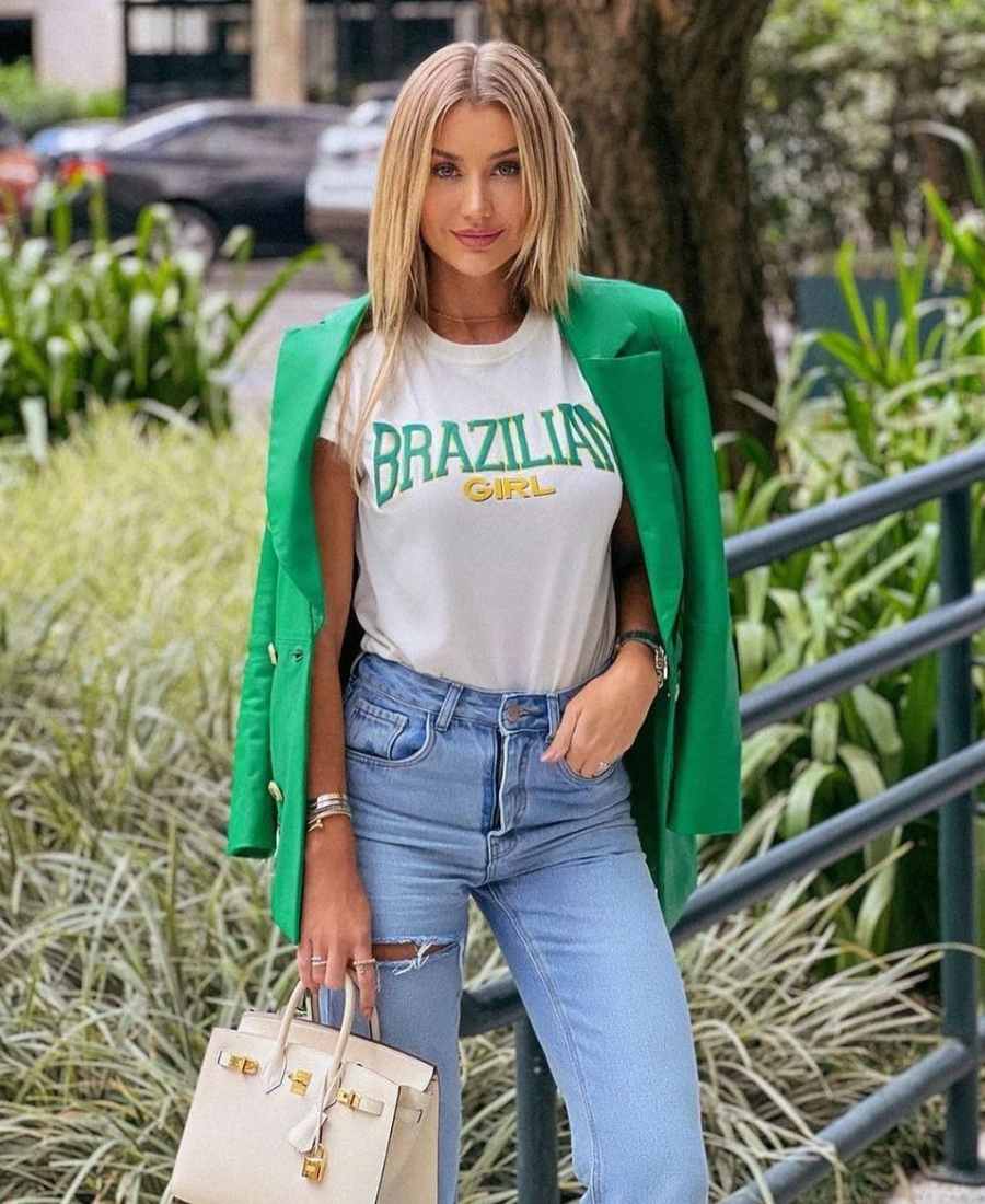 Imagem com fundo de natureza. No centro, mulher usando roupas Brazilcore: camiseta branca escrito "Brazilia Girl" + calça jeans + blazer verde + bolsa branca.