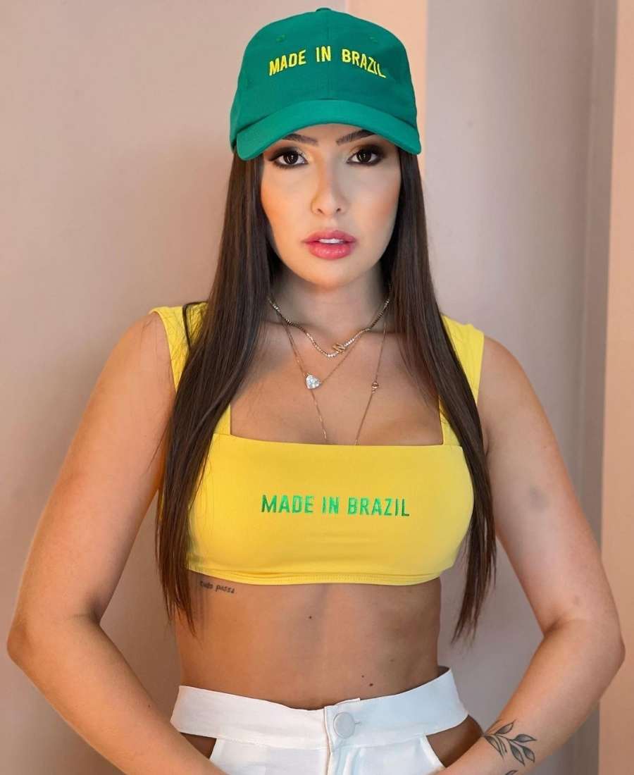 Imagem com fundo de parede. No centro, mulher usando roupas Brazilcore: top amarelo escrito "made in Brazil" + calça branca + boné verde.