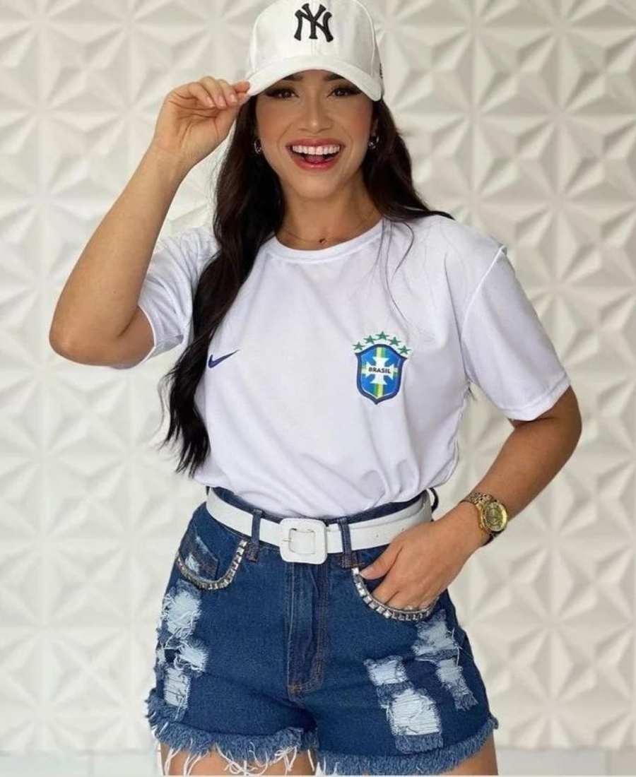 Imagem com fundo parede branca 3D. No centro, mulher usando roupas para a copa do mundo: camiseta branca do Brasil + short jeans com cinto branco+ boné branco.
