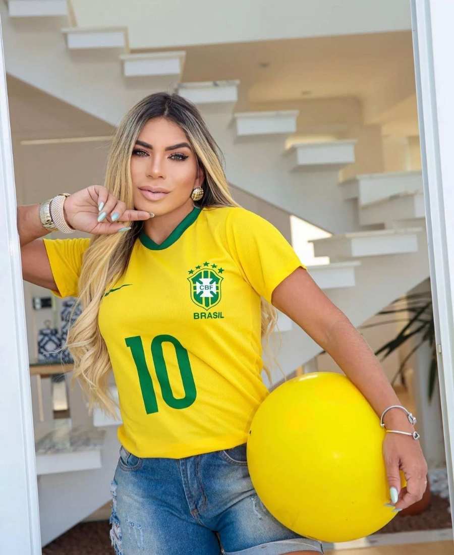 Imagem com fundo de cômodo de casa, com paredes e escada. No centro, mulher usando roupas Brazilcore: camiseta amarela do Brasil + short jeans, acessórios dourados e segurando bola amarela.