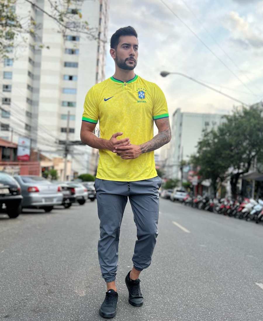 Imagem com fundo de rua com veículos e prédios. No centro, homem usando camiseta do Brasil: camiseta amarela + calça joger cinza + sapato preto.
