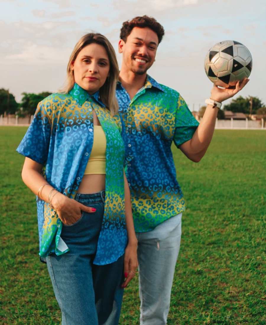 Imagem com fundo de campo de futebol. No centro, homem e mulher usando look para Copa do Mundo. Ambos usam camiseta verde, azul e amarela com estampa de onça.