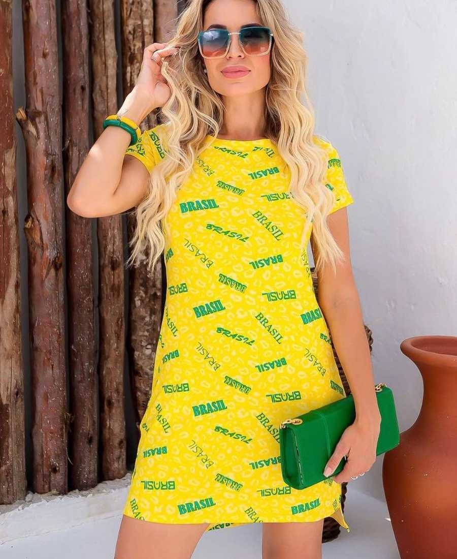 Imagem com fundo de parede e madeira. No centro, mulher usando roupa copa do mundo: vestido amarelo com estampa de oncinha escrito "Brasil" + bolsa verde + pulseiras verde e amarelo + óculos de sol.