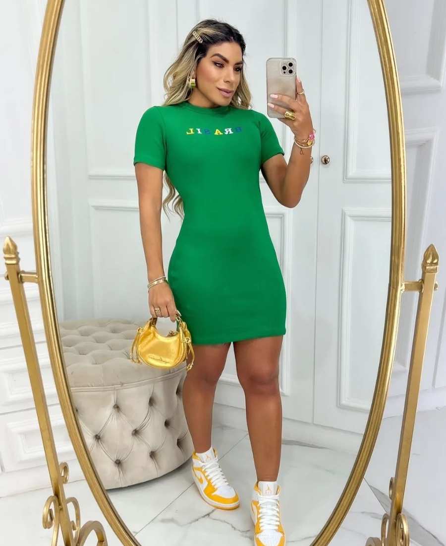 Imagem com fundo de parede, estofado e espelho. No centro, mulher usando roupa copa do mundo: vestido verde do Brasil + tênis branco e amarelo + bolsa dourada.