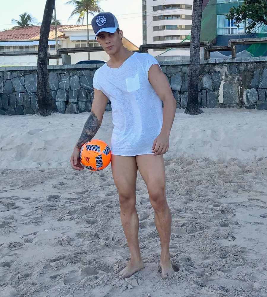 Imagem em fundo de areia da praia. Tiago Ramos em 2017, usando camisa branca, boné e segurando bola .