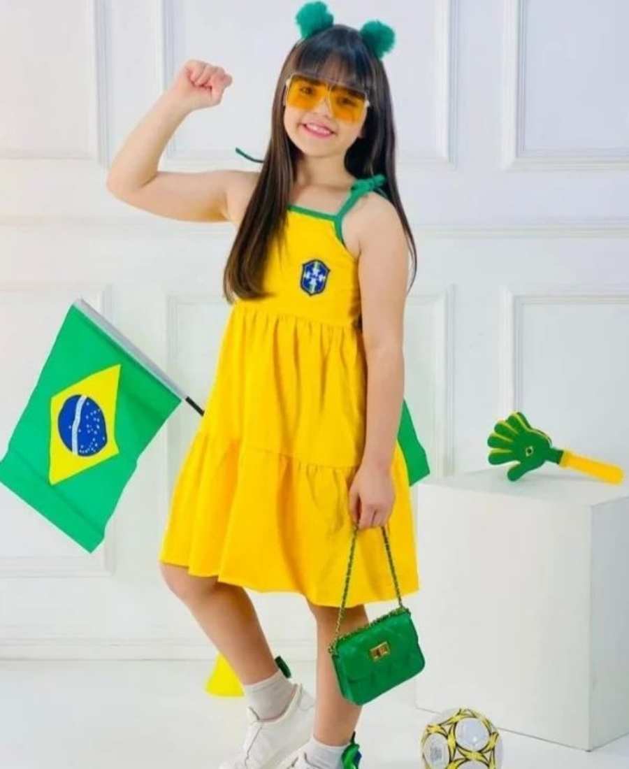 Imagem com fundo de parede branca. No centro, menina criança usando roupa para Copa do Mundo: vestido amarelo do Brasil + bolsa verde + tiara de pompom verde.
