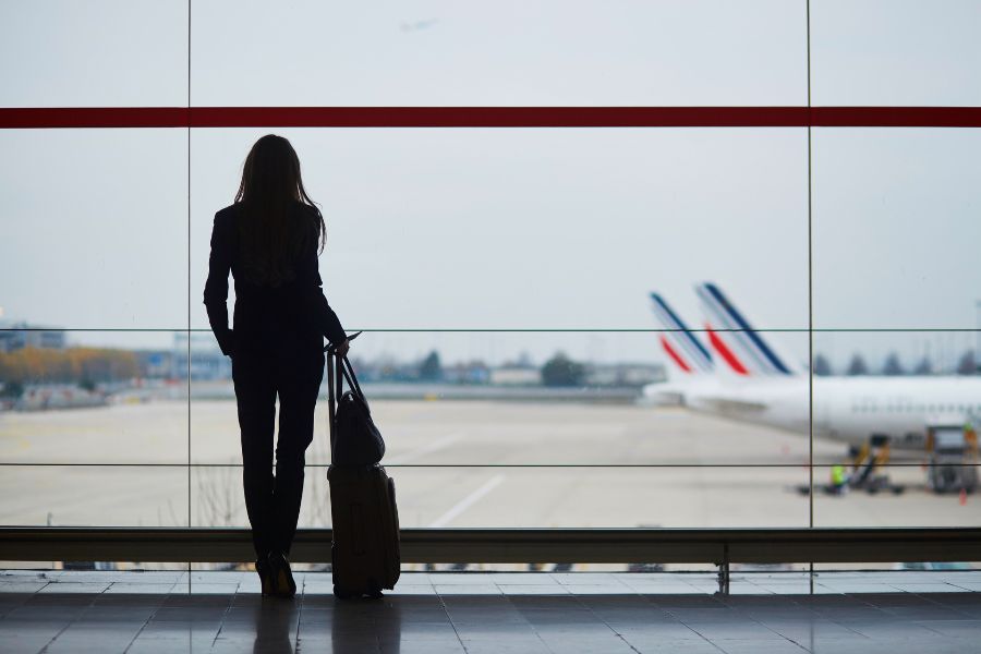 Mulher segurando malas e olhando pela janela de um aeroporto, qual a melhor forma de comprar passagens aéreas baratas