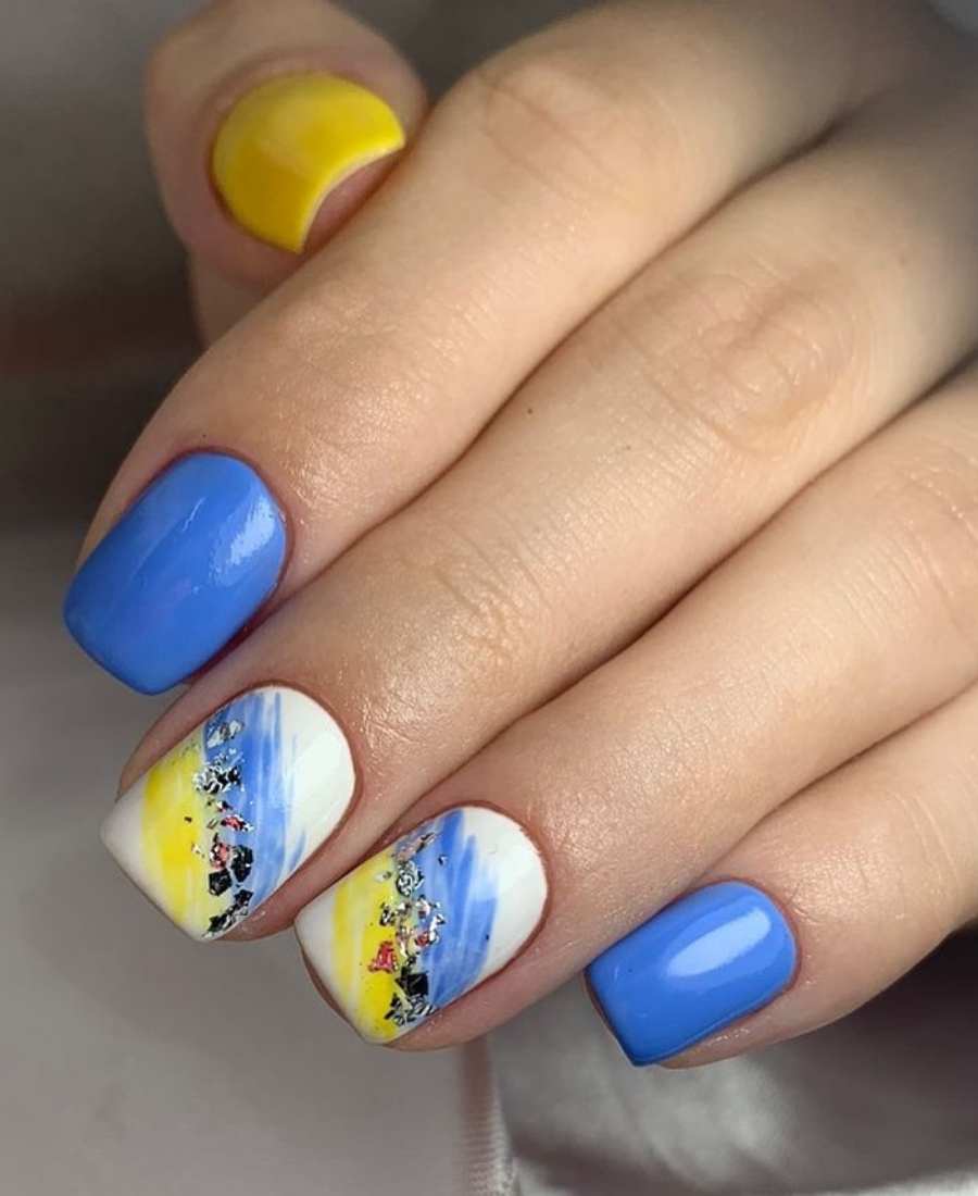 Imagem de mão com unhas decoradas para a Copa do Mundo 2022. Cores usadas: azul e amarelo. Polegar, amarelo. Indicador, azul-claro. Médio e anelar, branco com amarelo e azul com brilho dourado. Mínimo, azul.
