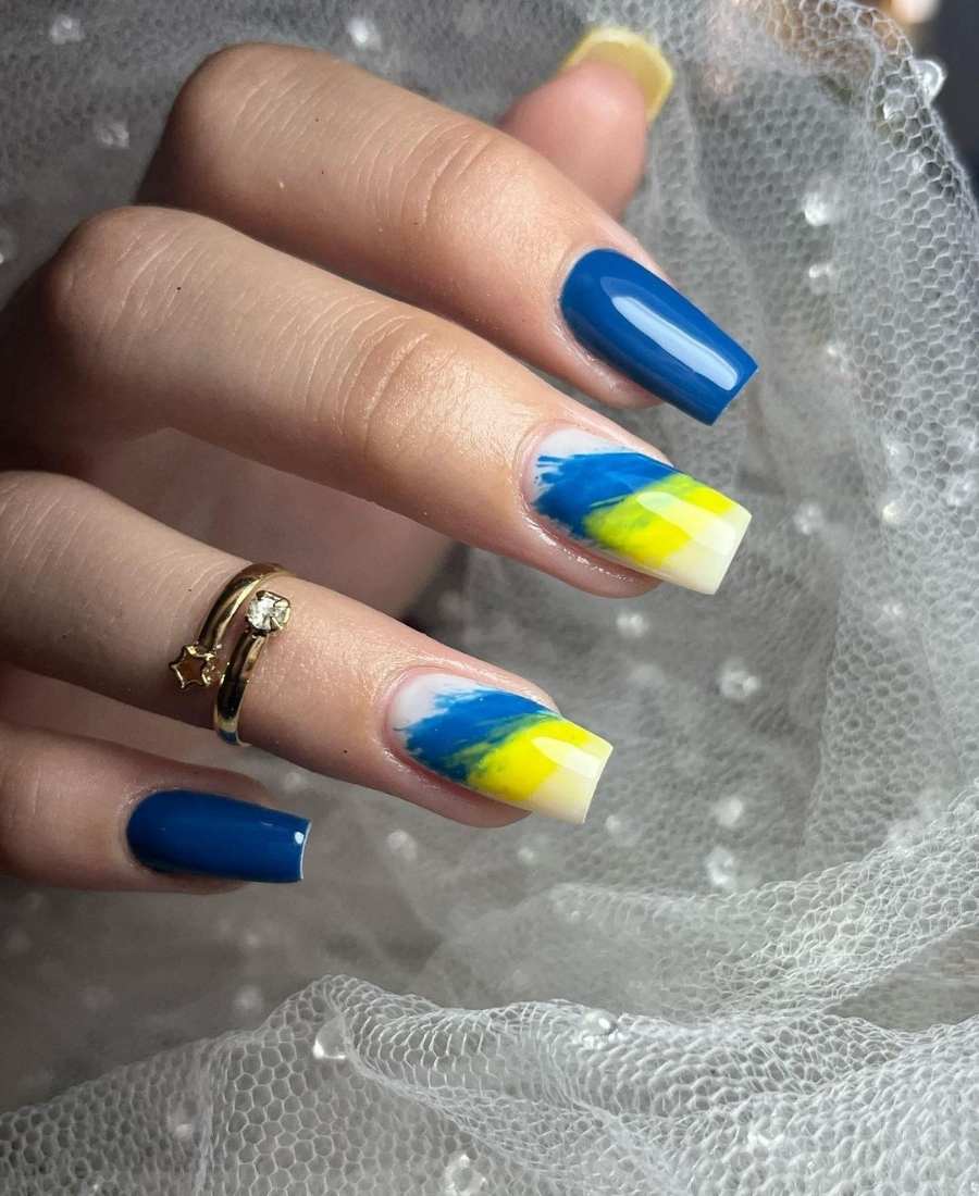 Imagem de mão com unhas decoradas para a Copa do Mundo 2022. Cores usadas: azul e amarelo. Polegar, amarelo. Indicador e mínimo, azul. Médio e anelar, branco com azul e amarelo. 