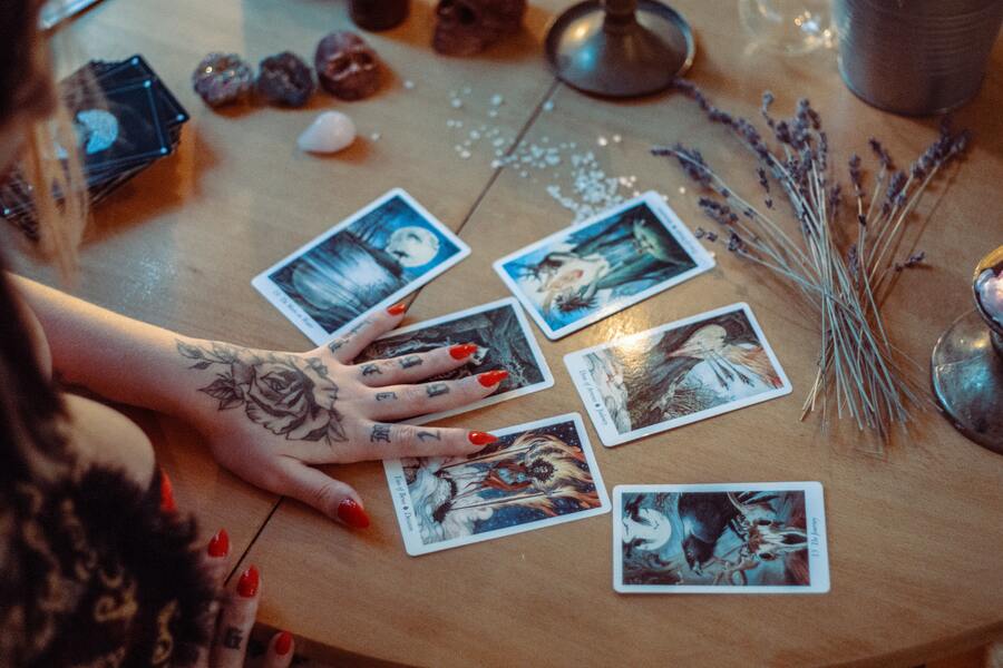 mulher com tatuagem na mão e unhas vermelhas tirando tarot em uma mesa com 6 cartas e cristais