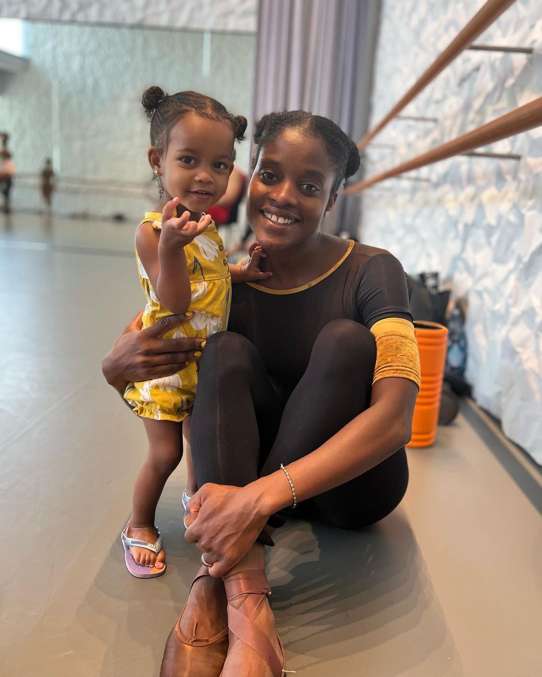 Imagem da bailarina Ingrid Silva e sua filha, Laura, sentadas em chão de sala de balé. 