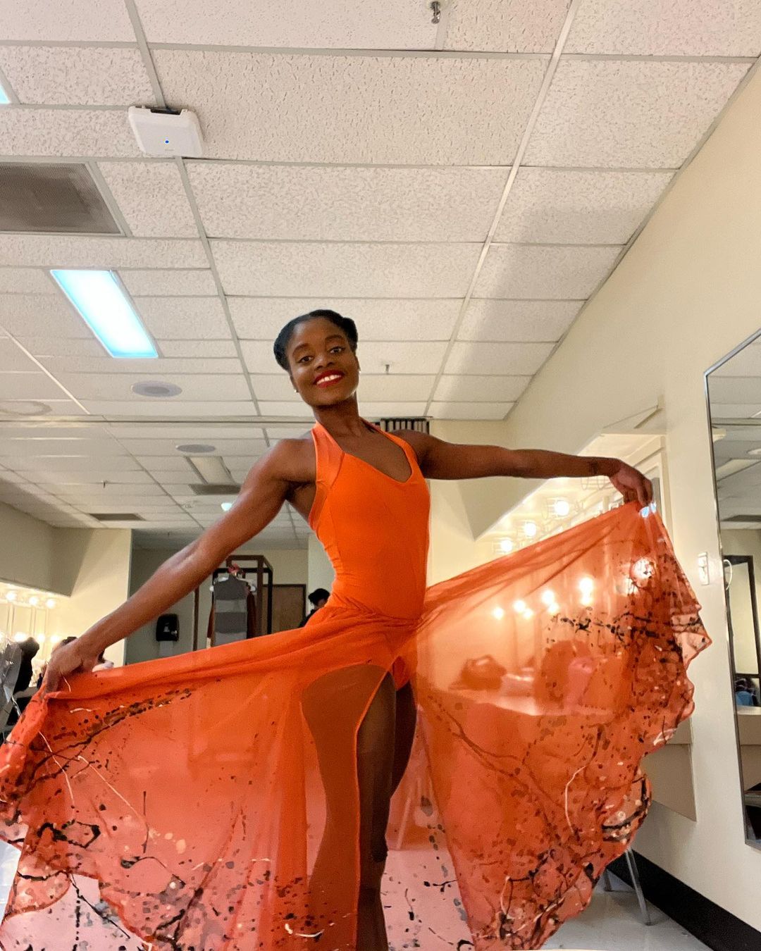 Imagem de bailarina brasileira Ingrid Silva, em local de ensaio, usando roupa de bailarina de cor laranja. 