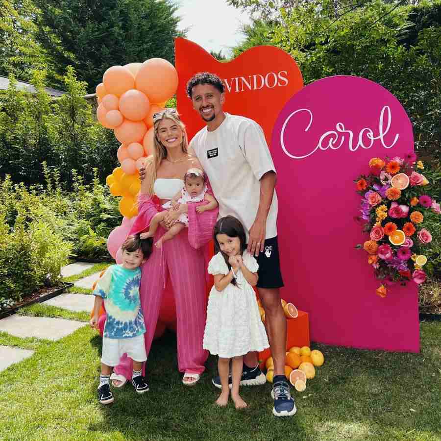 Foto de Marquinhos, zagueiro, com sua esposa Carol e seus filhos, no aniversário de 29 anos dela. Família posa de frente para painel decorativo com flores, plantas e frutas.