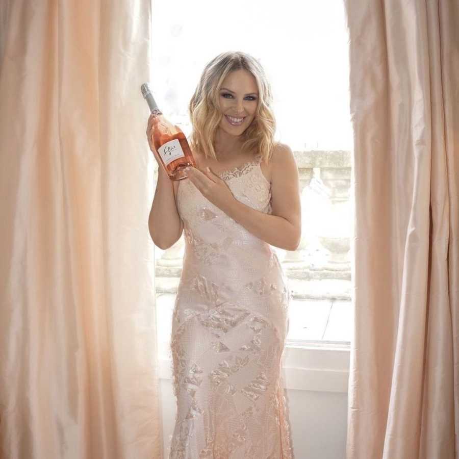 Imagem de Kylie Minogue, de pé em frente a janela, segurando garrafa de vinho. 