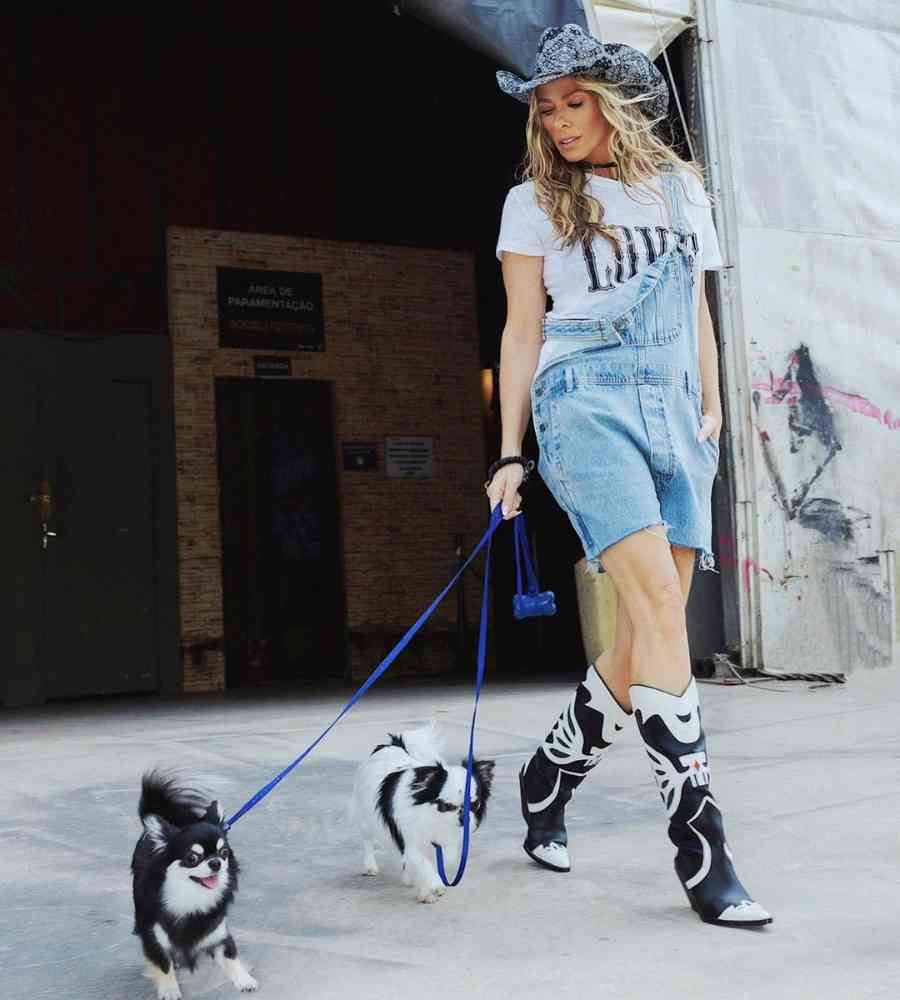 Foto de Adriane Galisteu usando macacão jeans + botas + chapéu, andando com 2 cachorros na coleira.